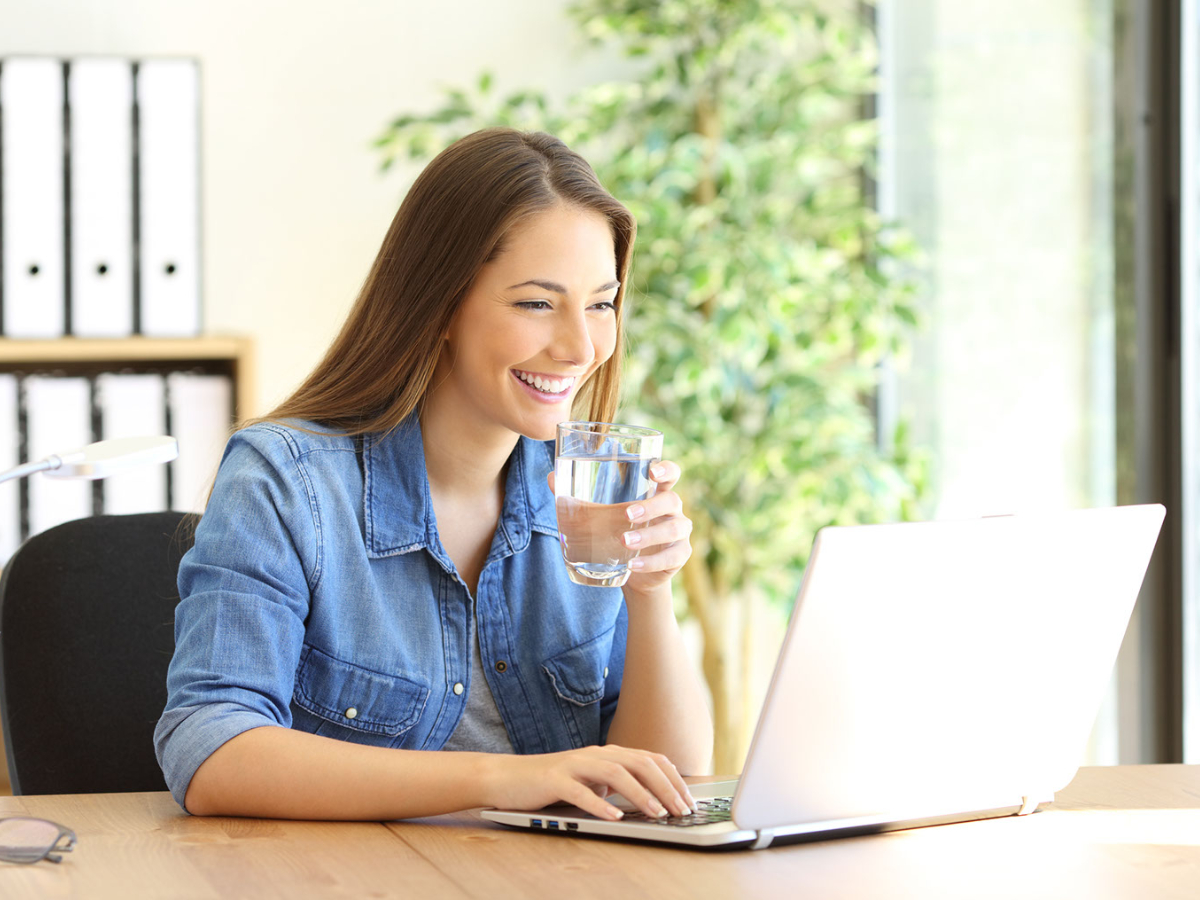 Mitarbeiter sind mit Wasser aus Wasserspender glücklich. Frau mit Wasserglas in der Hand, am Laptop sitzend