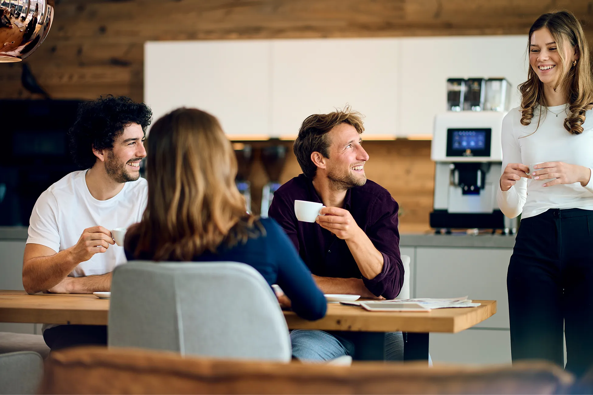 Kaffeemaschinen für Unternehmen. Vier Personen sitzen und stehen am Tisch, trinken Kaffee und unterhalten sich.